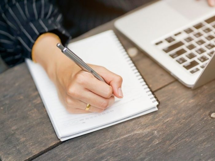Person writing notes in a notepad beside a laptop